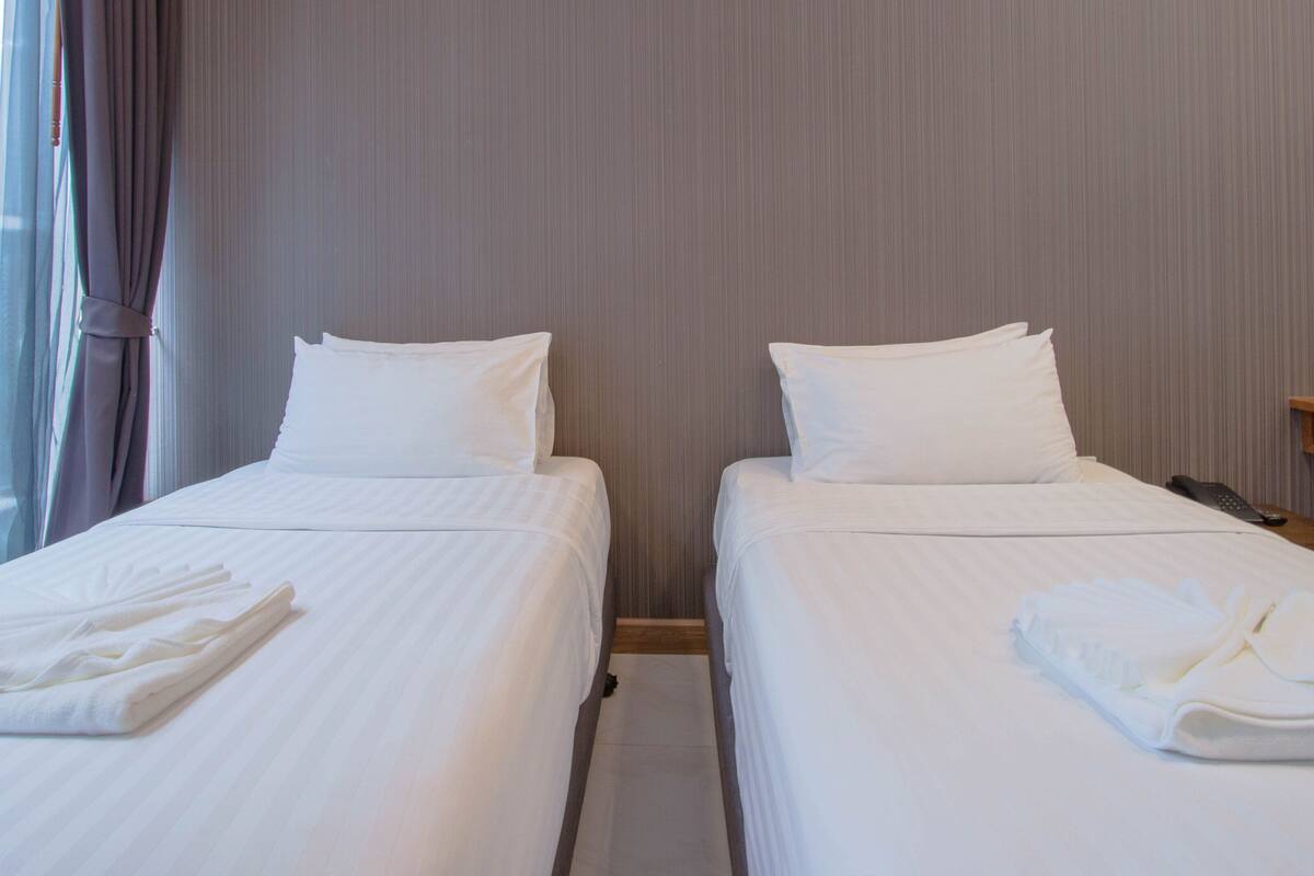 A hotel room with two beds, each with white linens and pillows, a wooden nightstand with a remote control, and a window with curtains.
