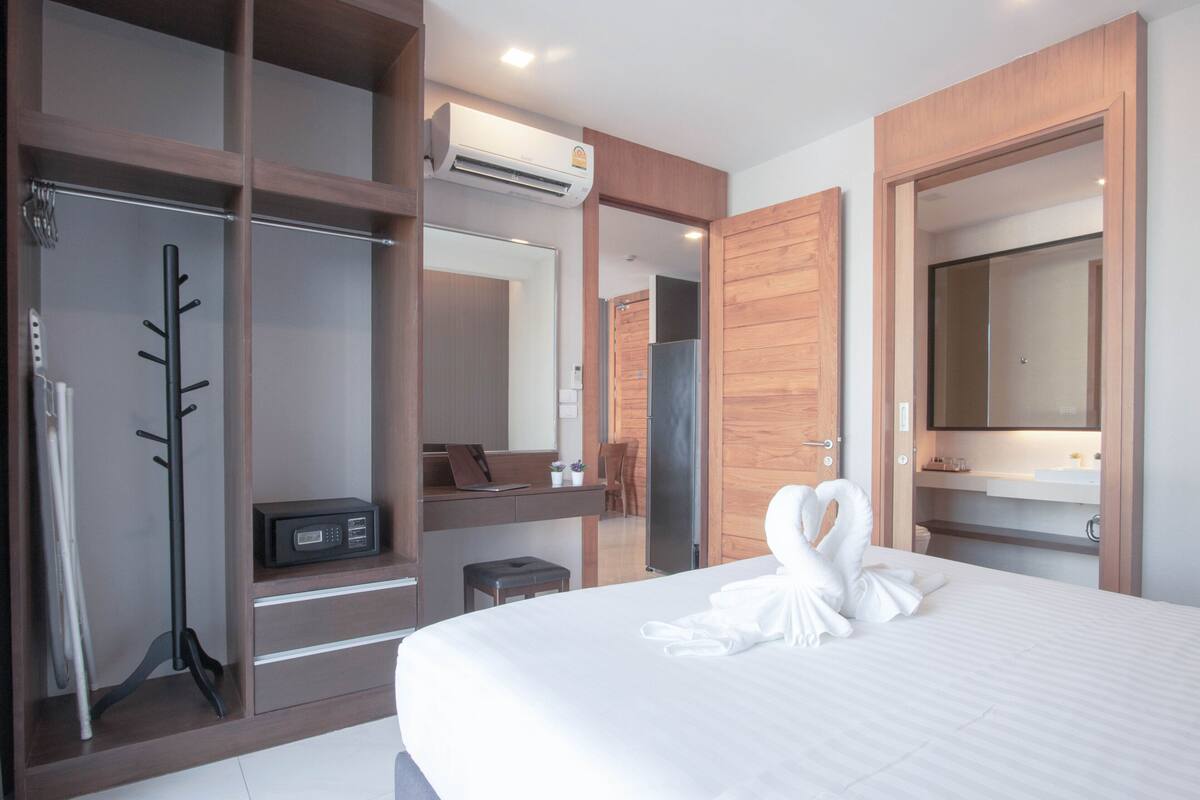 A hotel room with a large bed, a wooden wardrobe, a mirror, a desk with a laptop, and a coat rack.