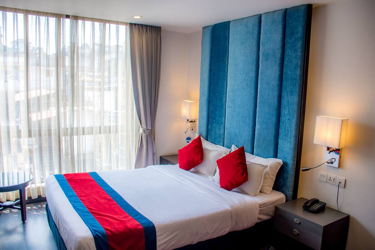 A hotel room with a large bed, two red pillows, a bedside table with a lamp, and a view of the city through sheer curtains.