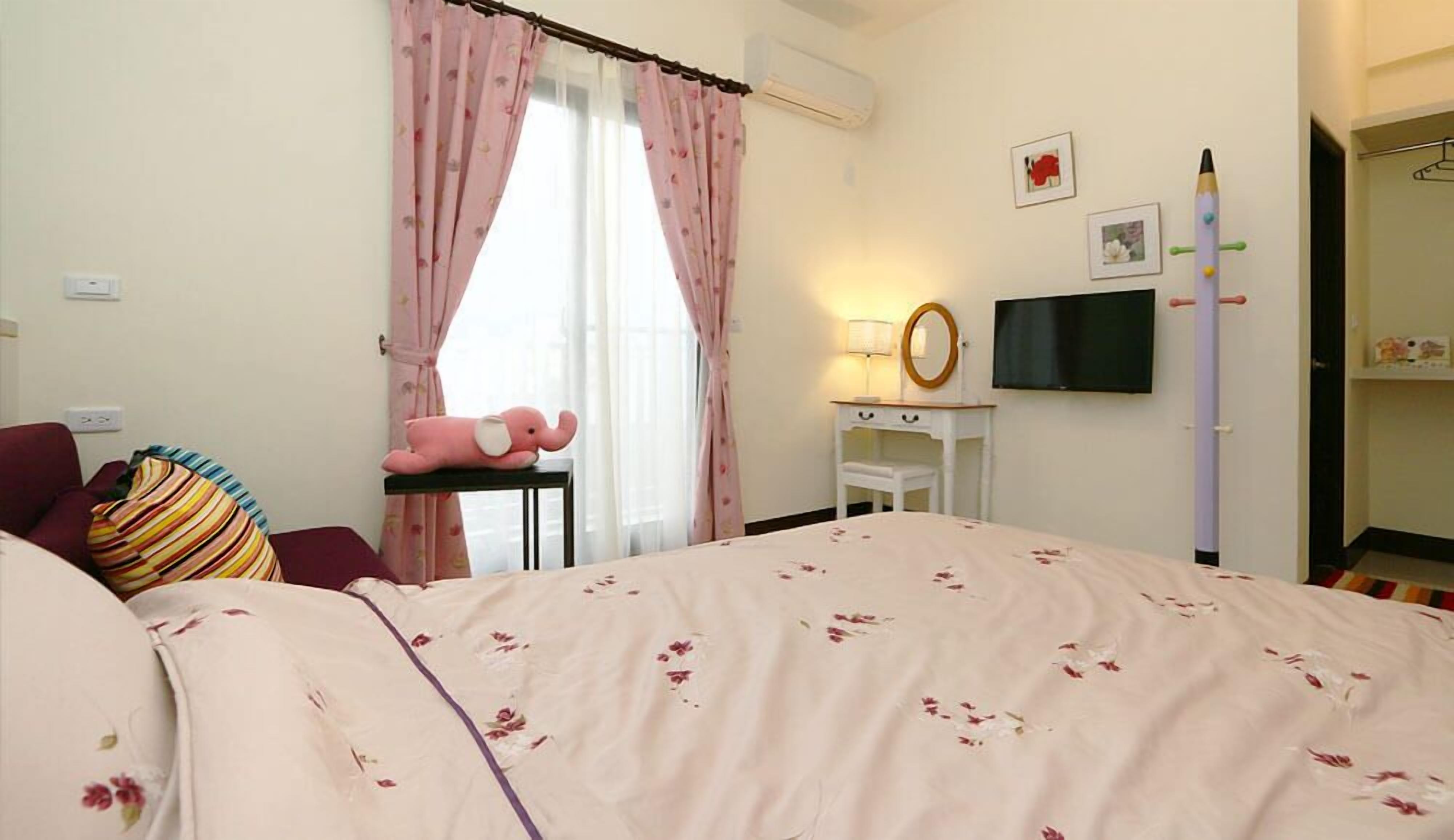 A bedroom with a bed, a desk, a chair, a television, and a pink elephant toy.