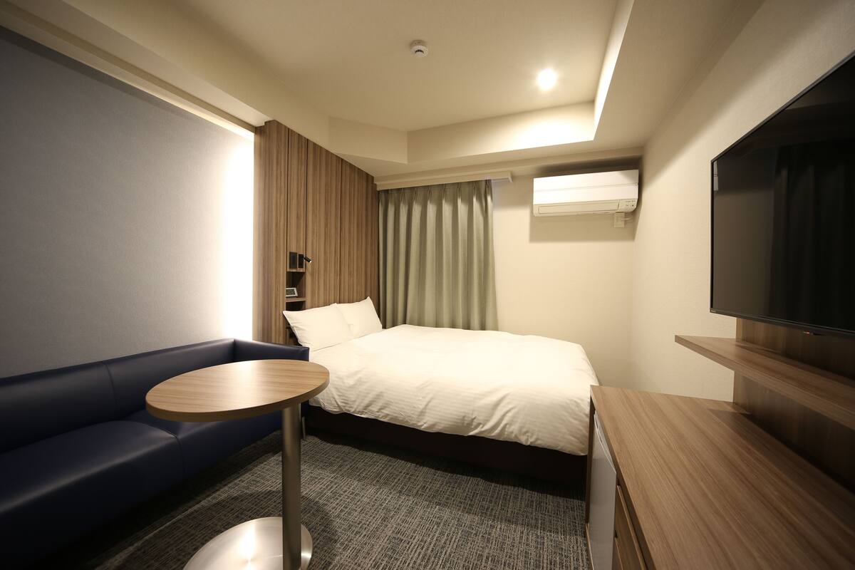 A hotel room with a bed, a sofa, a small round table, a TV, and an air conditioning unit.