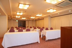 Een congreszaal met rijen tafels en stoelen, voorzien van plafondlampen en een airco.