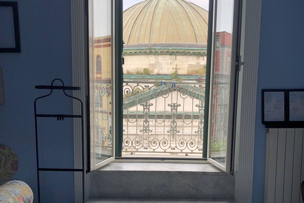 A room with a bed, a window with a view of a building with a dome, and a balcony with a railing.