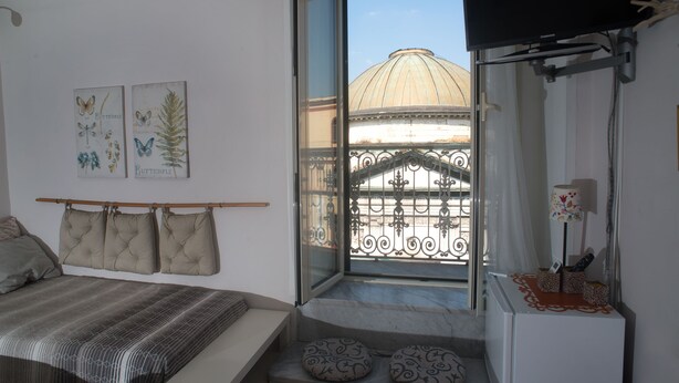 A bedroom with a bed, a bench, a window with a view of a dome and iron gate, and a TV mounted on the wall. - B&B Cuore di Napoli (Naples)
