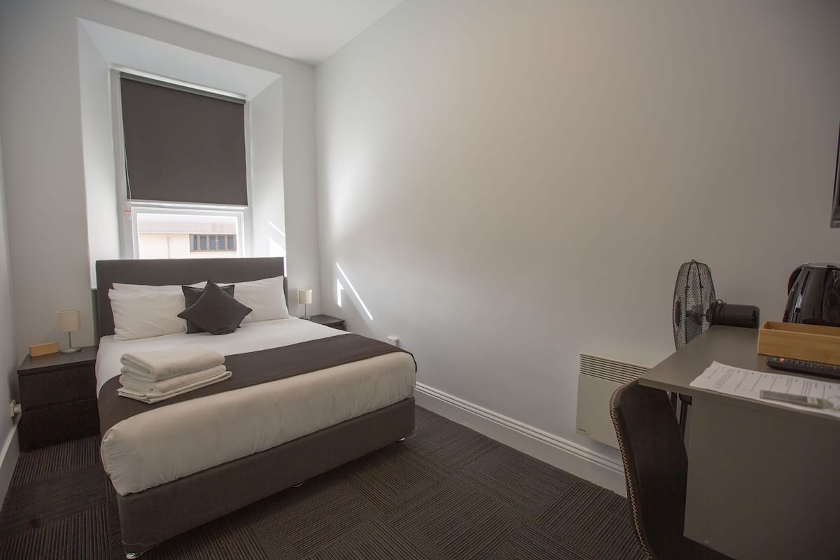 Quarto de hotel com uma cama, uma escrivaninha e uma cadeira.