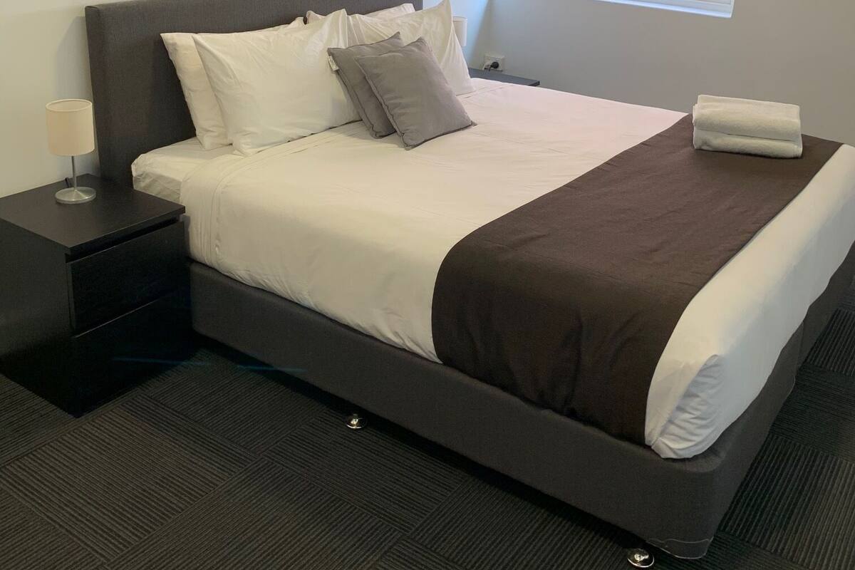 Uma cama bem arrumada com roupa de cama branca e castanha, uma mesa de cabeceira com abajur e uma janela com persianas.
