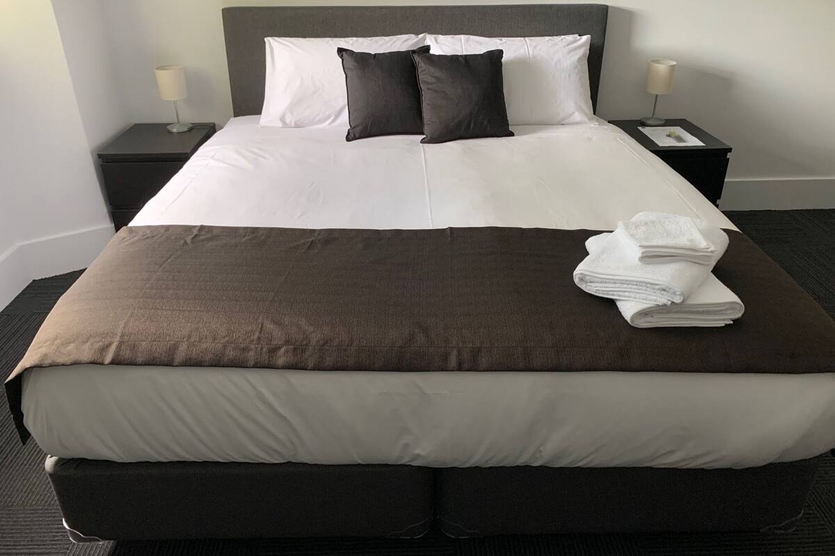 Cama bem arrumada com roupa de cama branca e marrom, dois travesseiros e uma toalha dobrada ao lado.