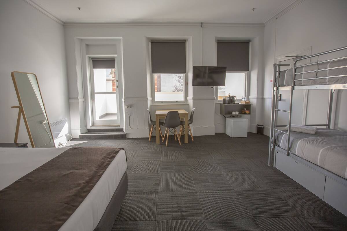 Quarto de hotel com cama, escrivaninha com cadeira, televisão e beliche.