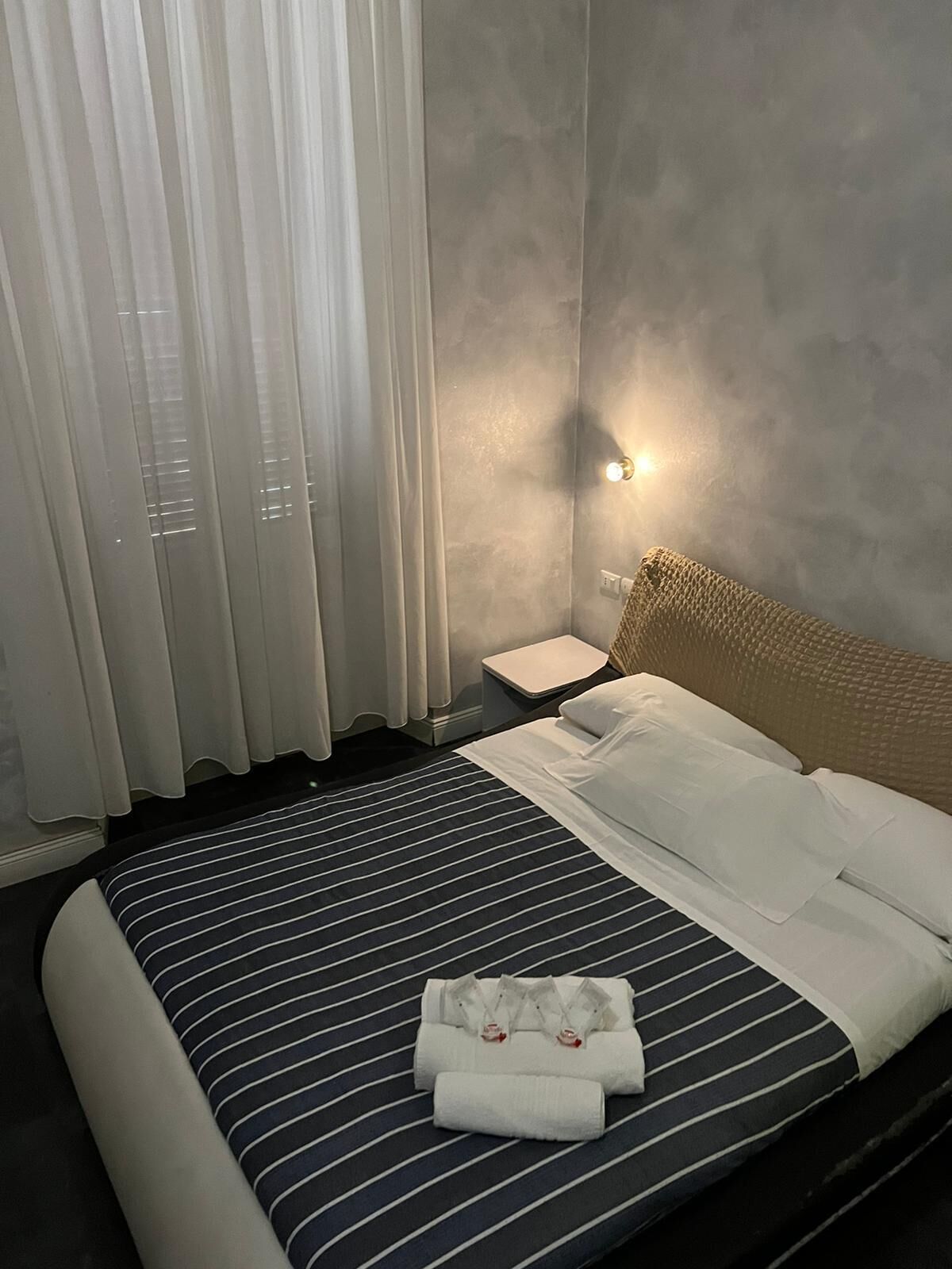 Una habitación de hotel con una cama, cortinas blancas, una mesita de noche y una lámpara de pared.