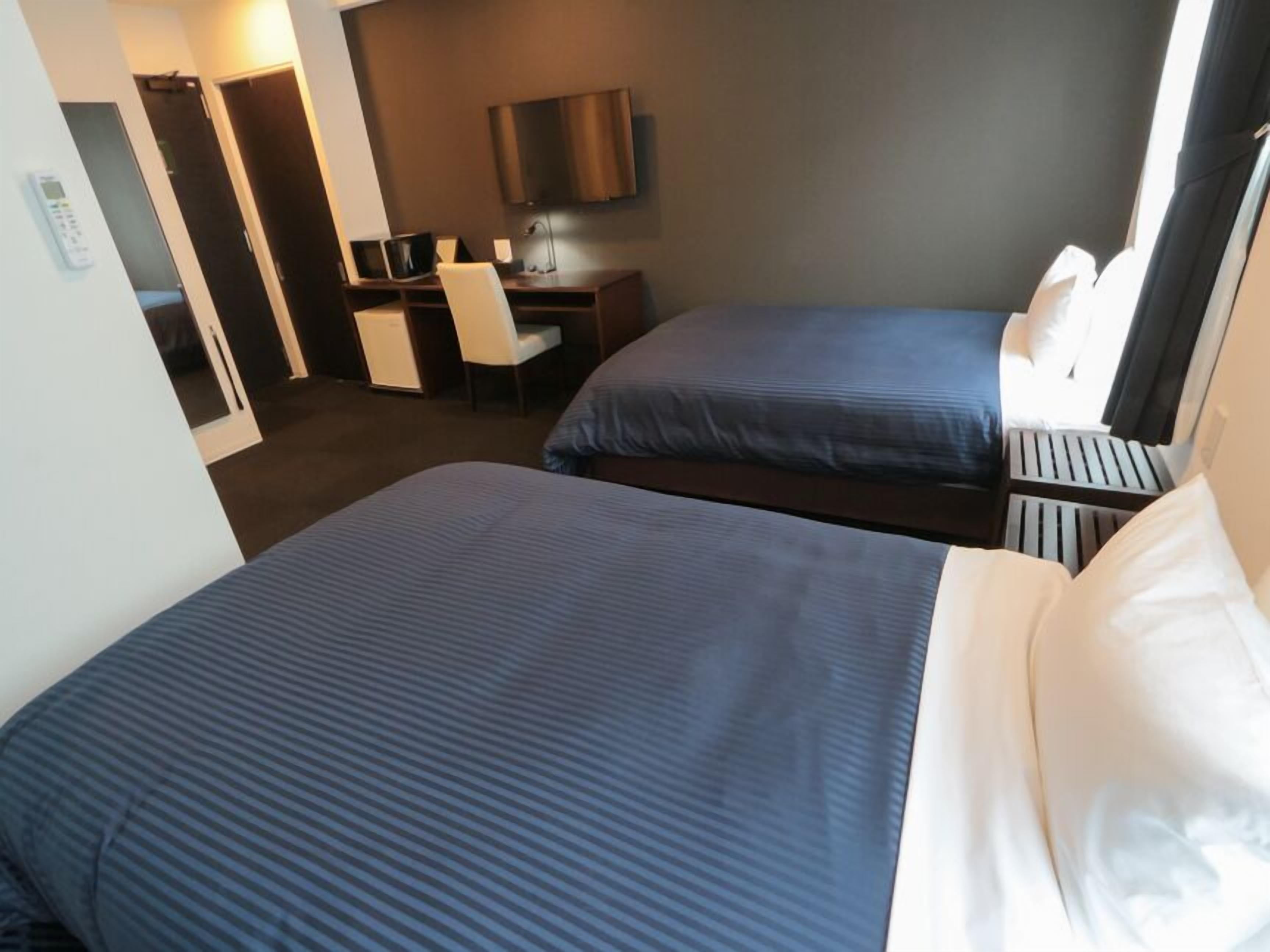 A hotel room with two beds, a desk, a chair, a TV, and a control panel.