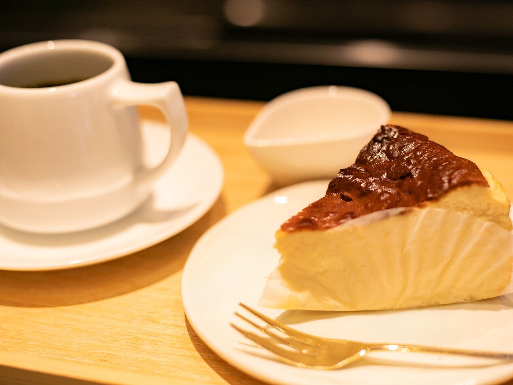 A slice of cheesecake with a chocolate topping on a white plate, accompanied by a cup of coffee and a small bowl on a wooden table.