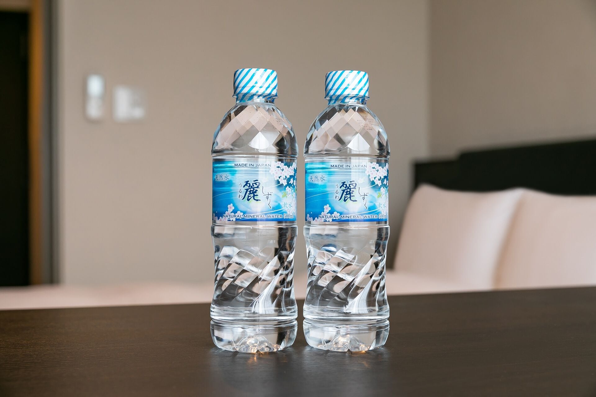 Dos botellas de agua con etiqueta azul y diseño de diamantes sobre una mesa de madera en una habitación con una cama y un almohadón blanco al fondo.