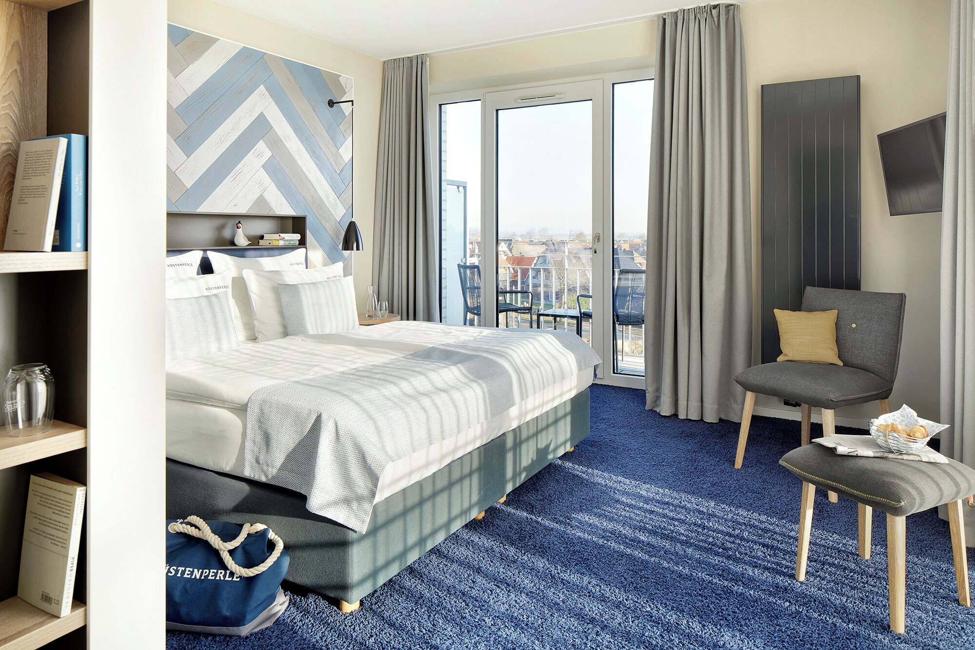 Ein Hotelzimmer mit einem Bett, einem Sessel, einem kleinen Tisch und Blick auf die Stadt durch große Fenster.