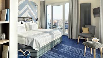 Ein Hotelzimmer mit einem Bett, einem Sessel, einem kleinen Tisch und Blick auf die Stadt durch große Fenster.