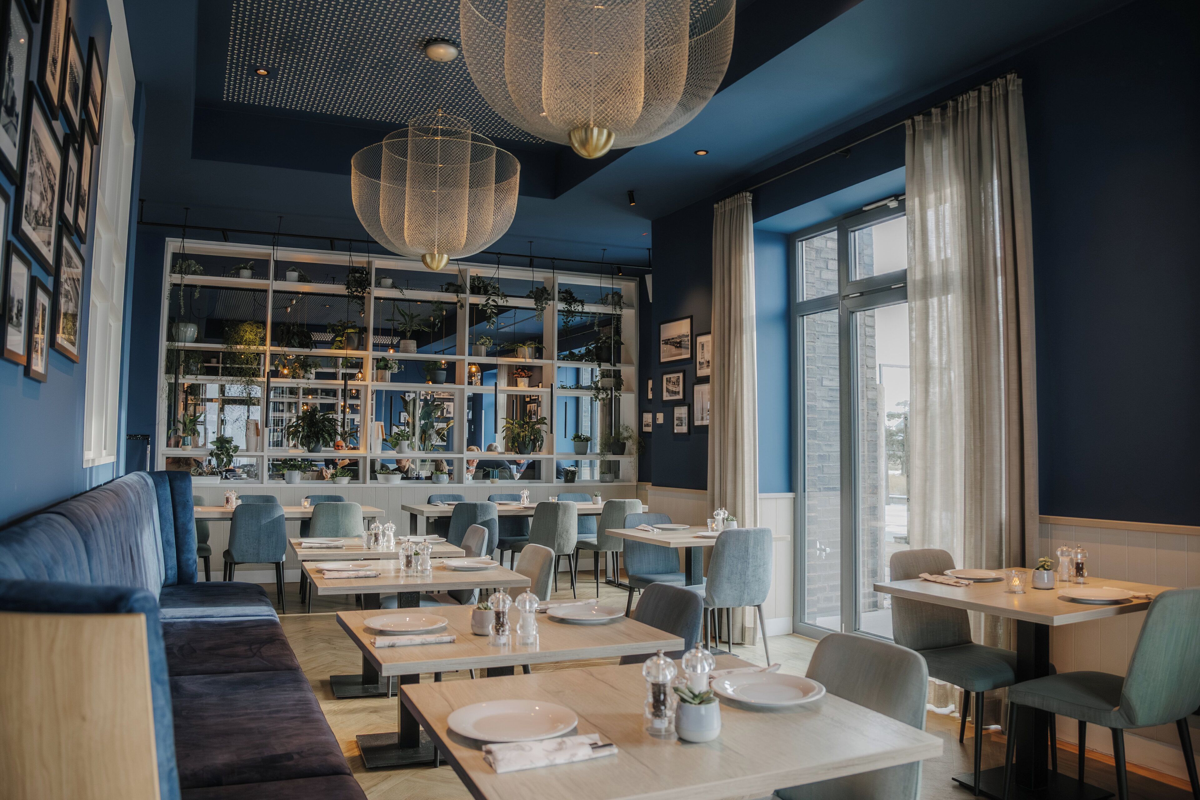 A modern restaurant with a blue interior, large windows, and a variety of seating options including tables and a sofa.