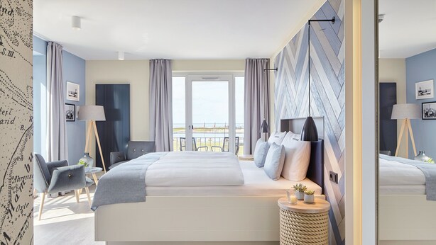 A modern hotel room with a large bed, a seating area with a chair and a small table, and a view of the beach through the window. - Küstenperle Strandhotel & Spa (Büsum)
