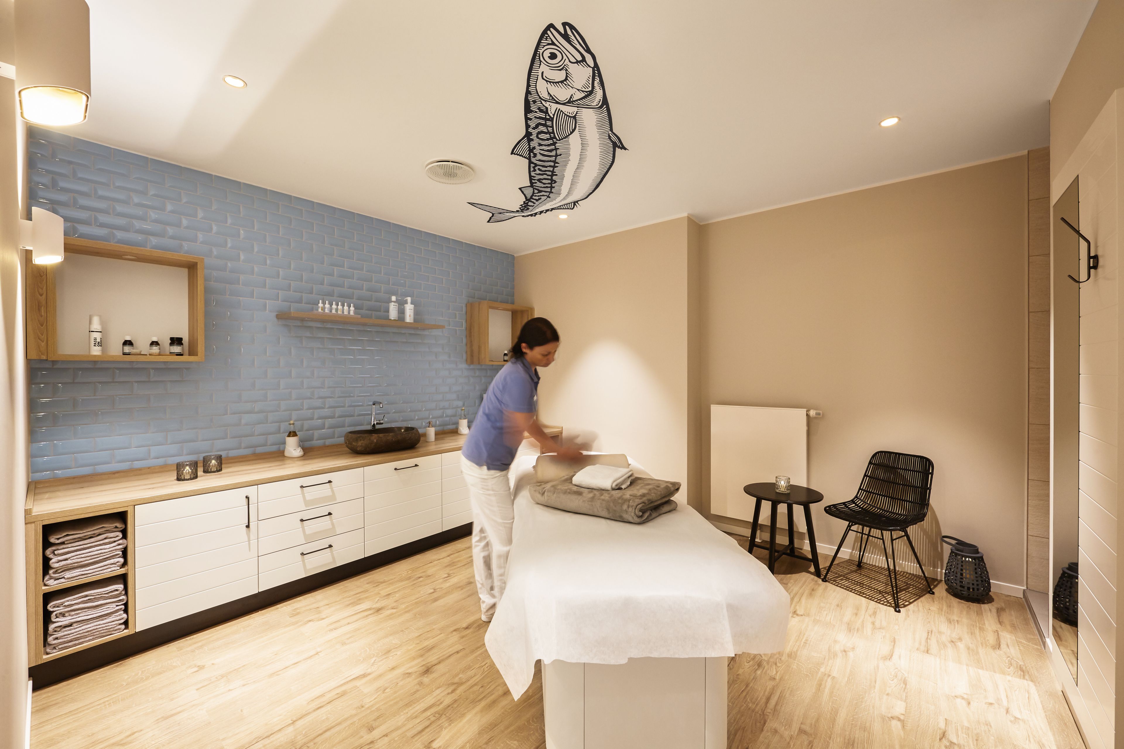 A room with a massage table, a blue-tiled wall, a wooden cabinet, and a fish decal on the ceiling.