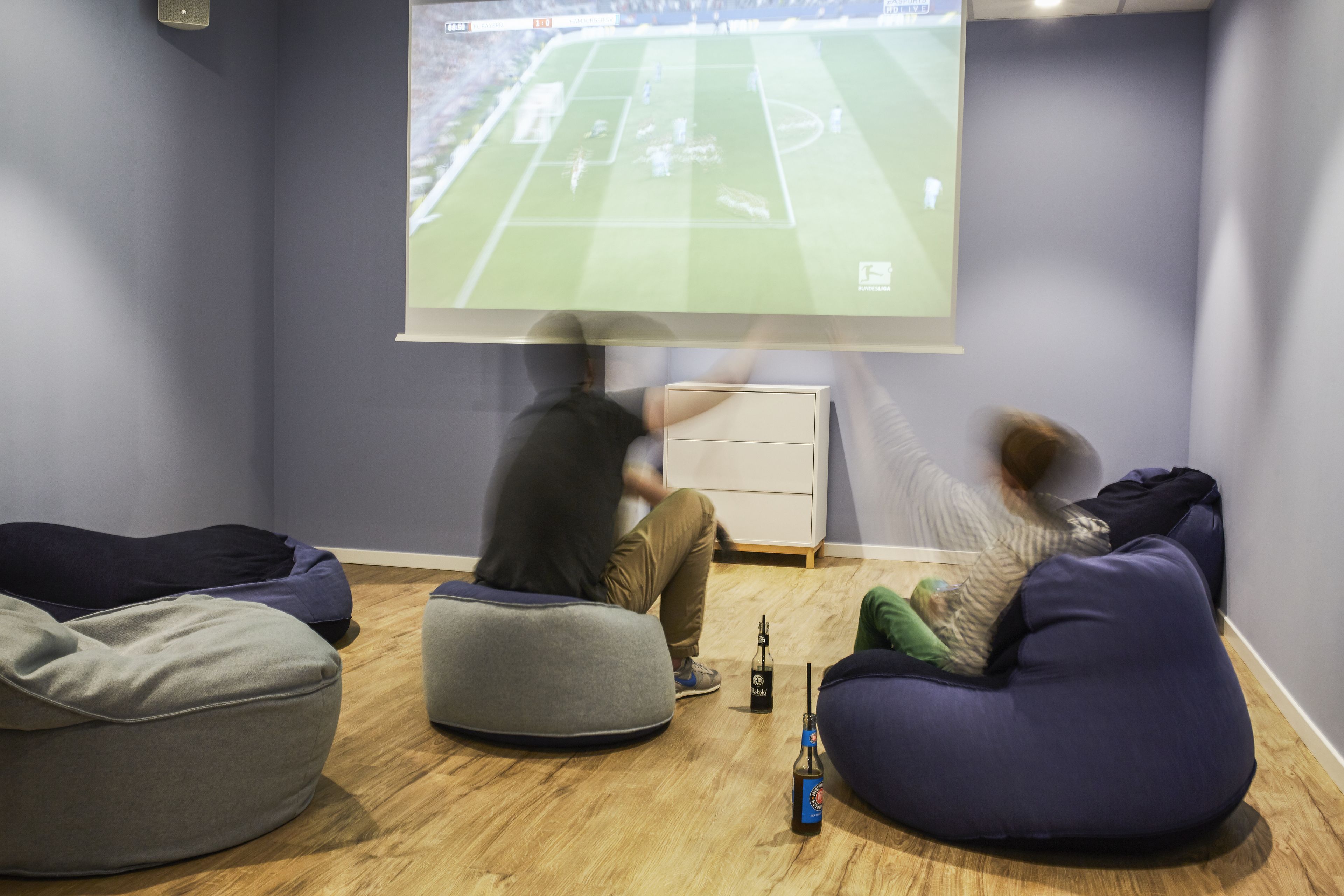 A room with a large screen displaying a sports game, bean bags, and a bottle on the floor.