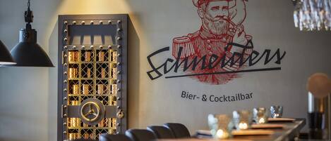 A bar area with a sign reading "Schneider Bier- & Cocktailbar" and a decorative wall with a figure and birds.