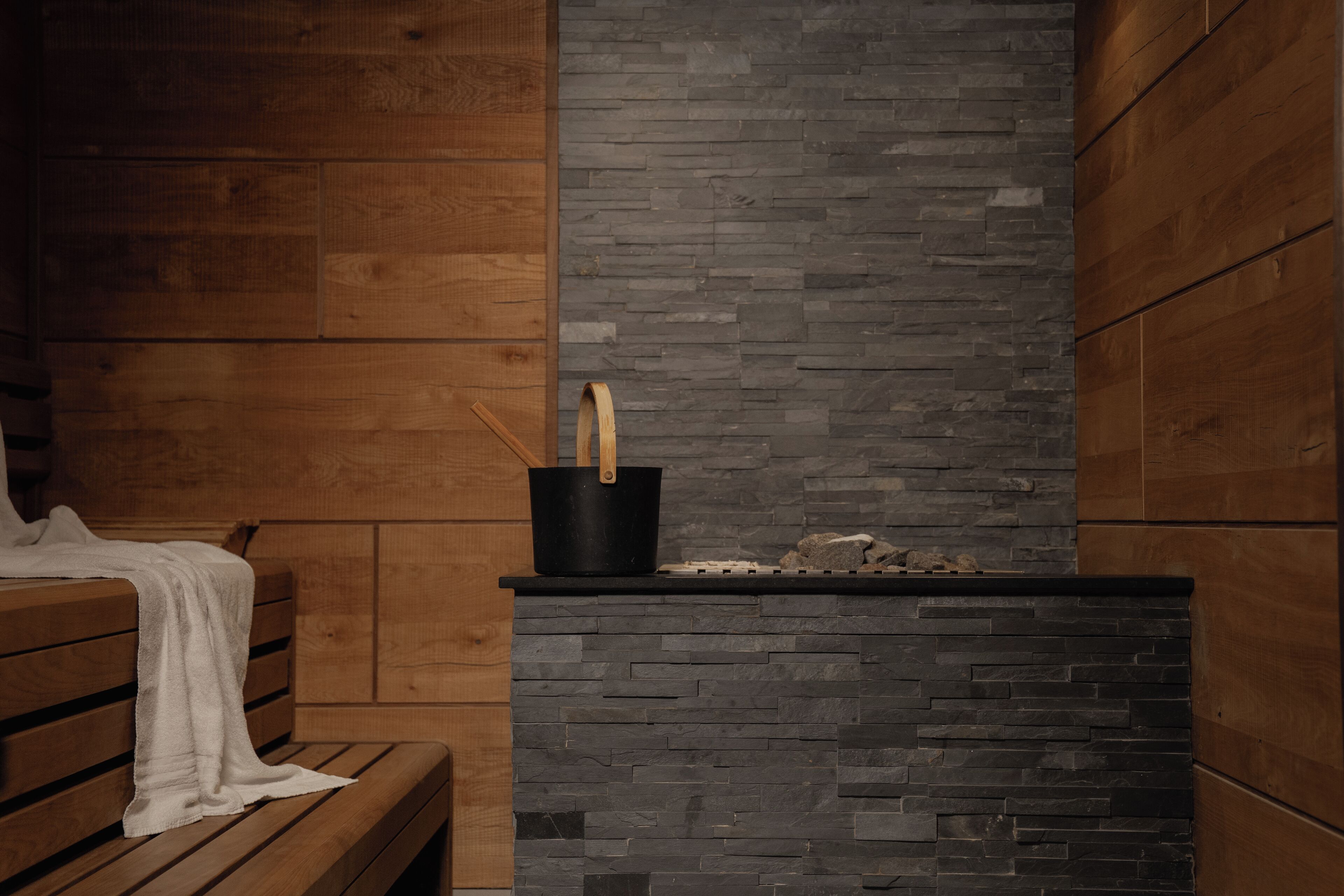 A wooden bench with a white towel, a black bucket with a wooden handle, and a stone wall.
