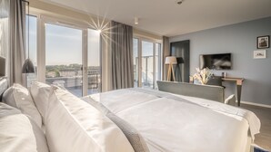 A hotel room with a large bed, a desk, a TV, and a view of the city through large windows. - Küstenperle Strandhotel & Spa (Büsum)