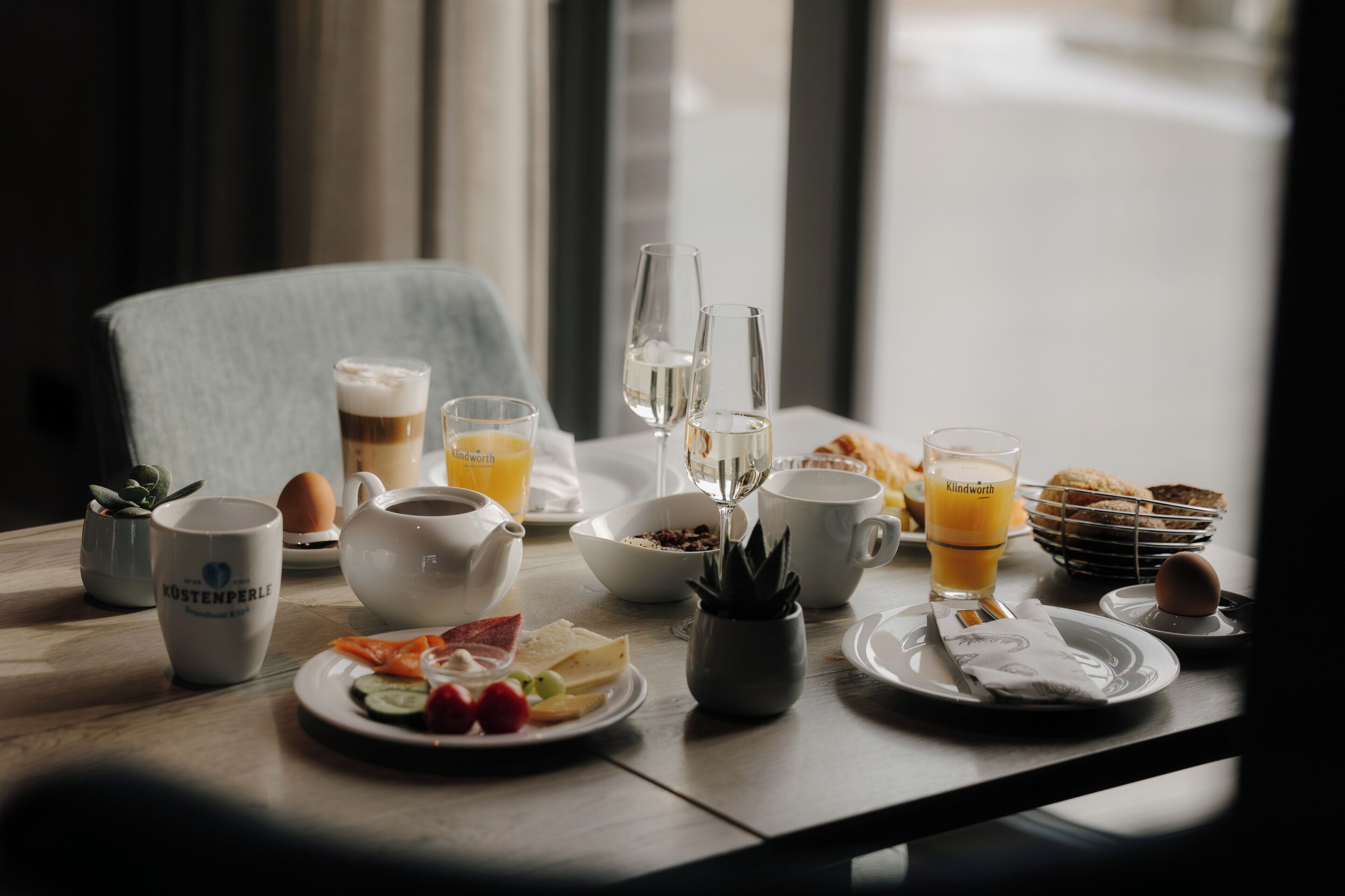 Ein Frühstückstisch mit verschiedenen Speisen, Getränken und einer Tasse mit der Aufschrift 'KÖSTENPERLE'.