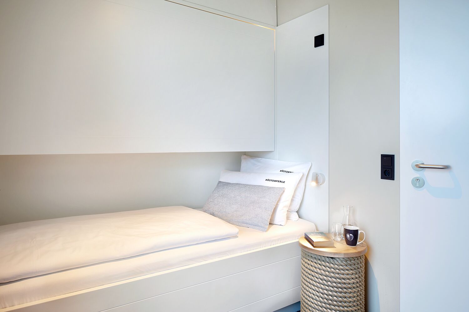 A small, neatly arranged bedroom with a bed, pillows, a bedside table, and a waste basket.