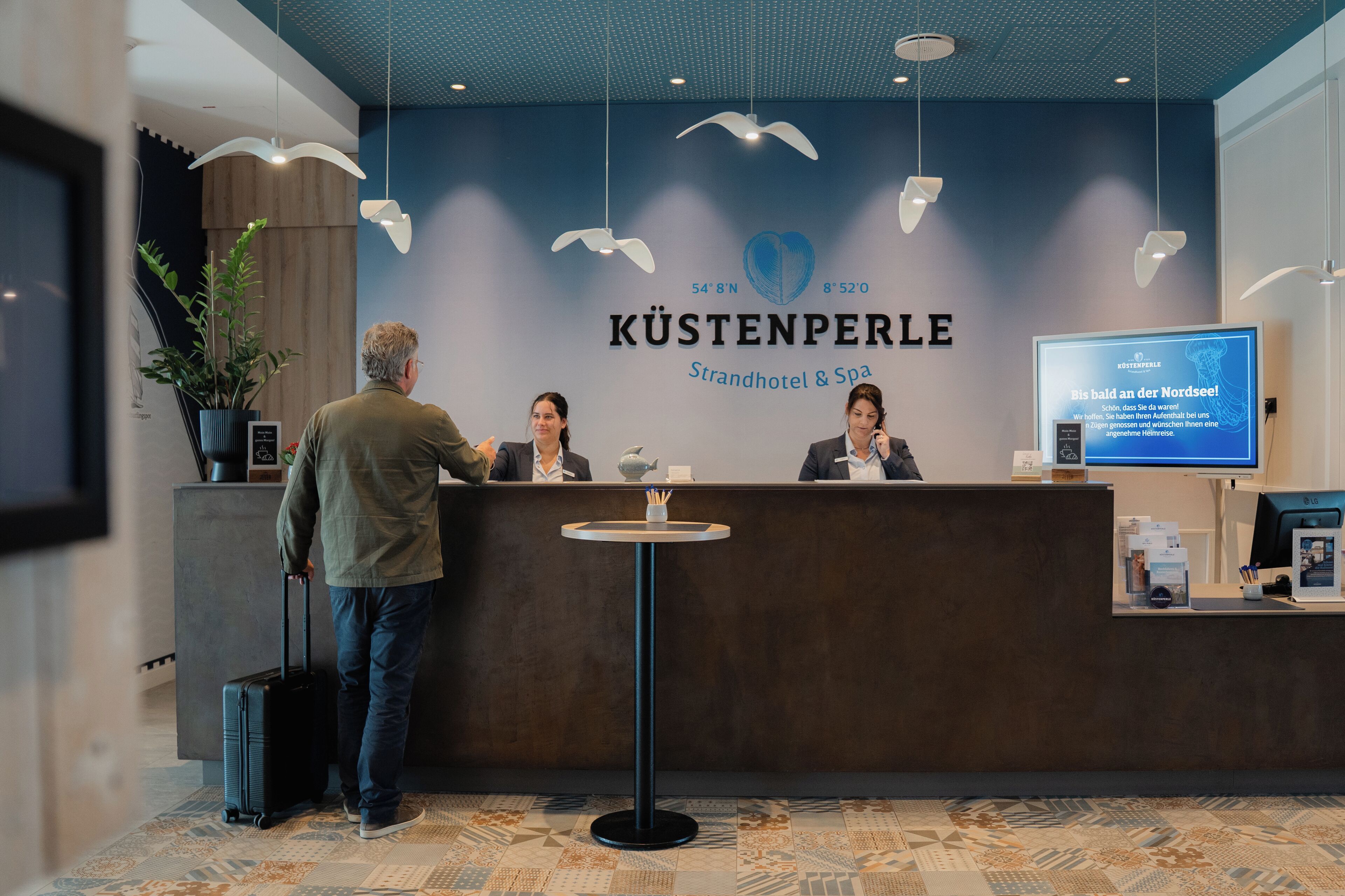 Un mostrador de recepción en KÜSTENPERLE Strandhotel & Spa con un diseño moderno, una pantalla digital que muestra información y un suelo con estampado.