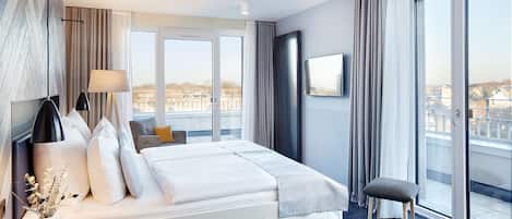 A modern hotel room with a large bed, a small seating area, a TV, and a view of the city.