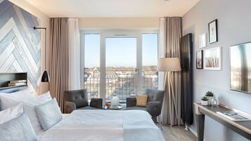 Ein modernes Hotelzimmer mit einem großen Bett, einer Sitzecke mit zwei Sesseln, einem Schreibtisch und einem großen Fenster mit Blick auf die Stadt.