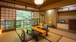 Une pièce de style japonais traditionnel, avec un sol recouvert de tatamis, une table basse agrémentée d’un chemin de table, un banc rembourré, une chaise et une porte coulissante donnant sur un paysage verdoyant.
