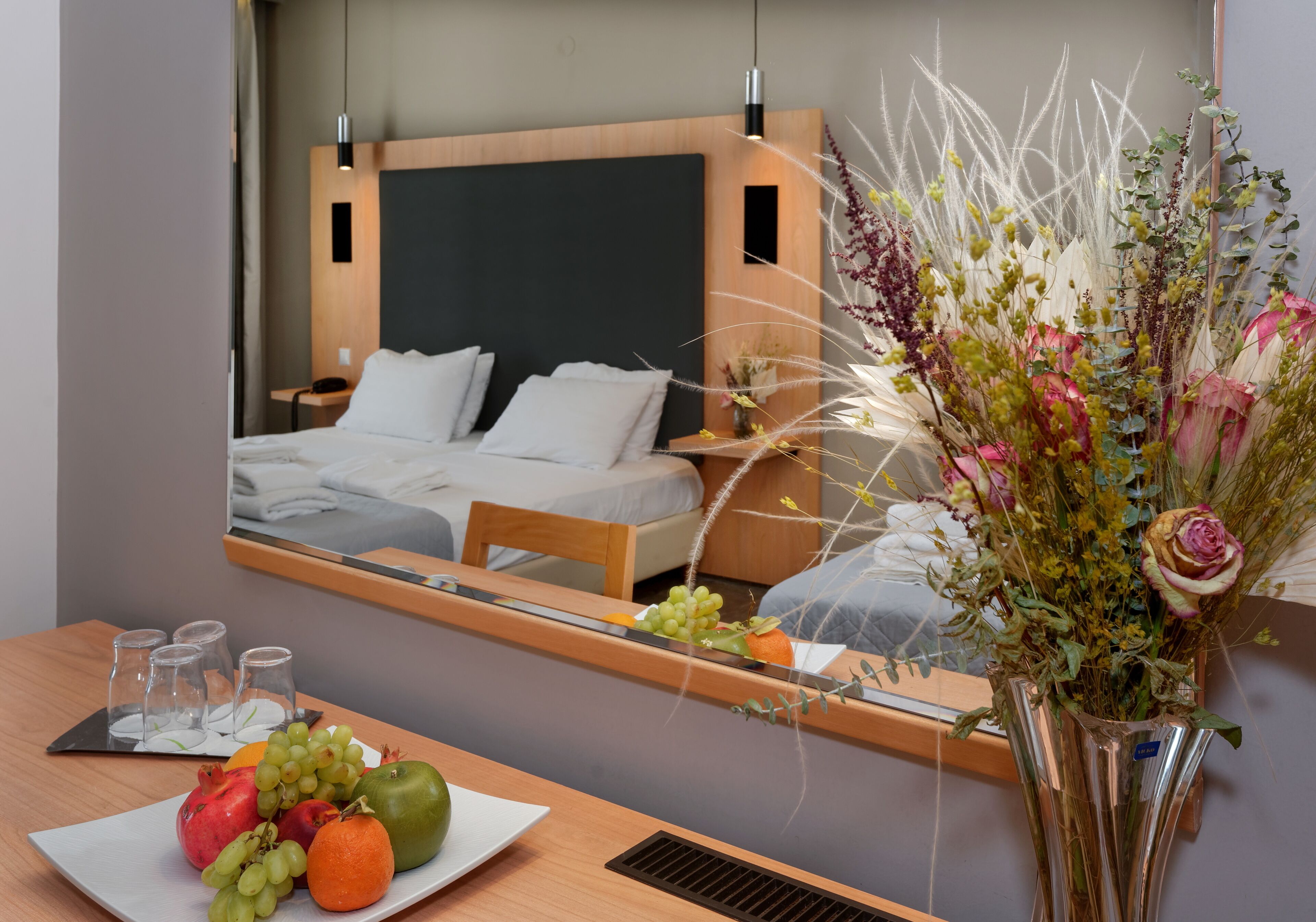 A hotel room with a bed, bedside table, and a mirror reflecting the room. There is a vase with flowers on the table.
