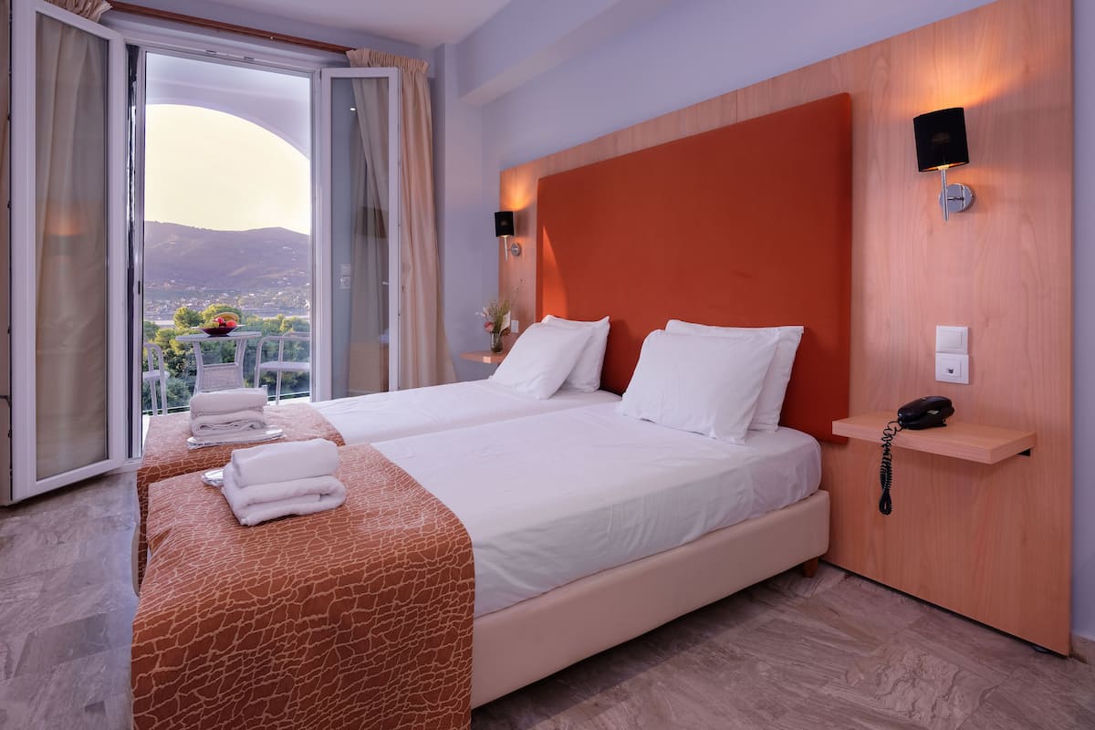 Ein Hotelzimmer mit zwei Betten, Blick ins Freie, einem hölzernen Kopfteil, einem Nachttisch mit Telefon und einer Glastür zum Balkon.