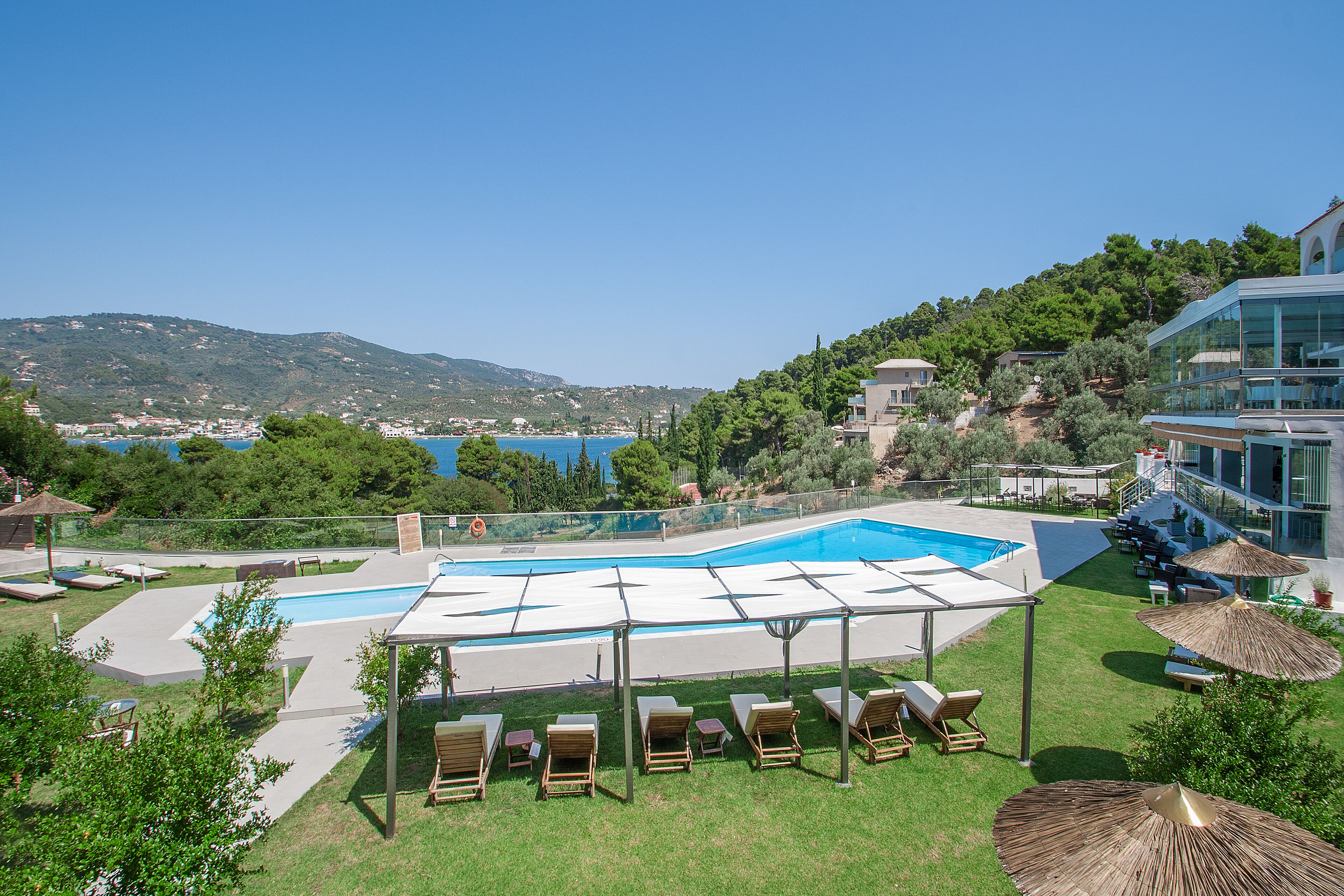 A pool area with lounge chairs and umbrellas, surrounded by greenery and overlooking a body of water.