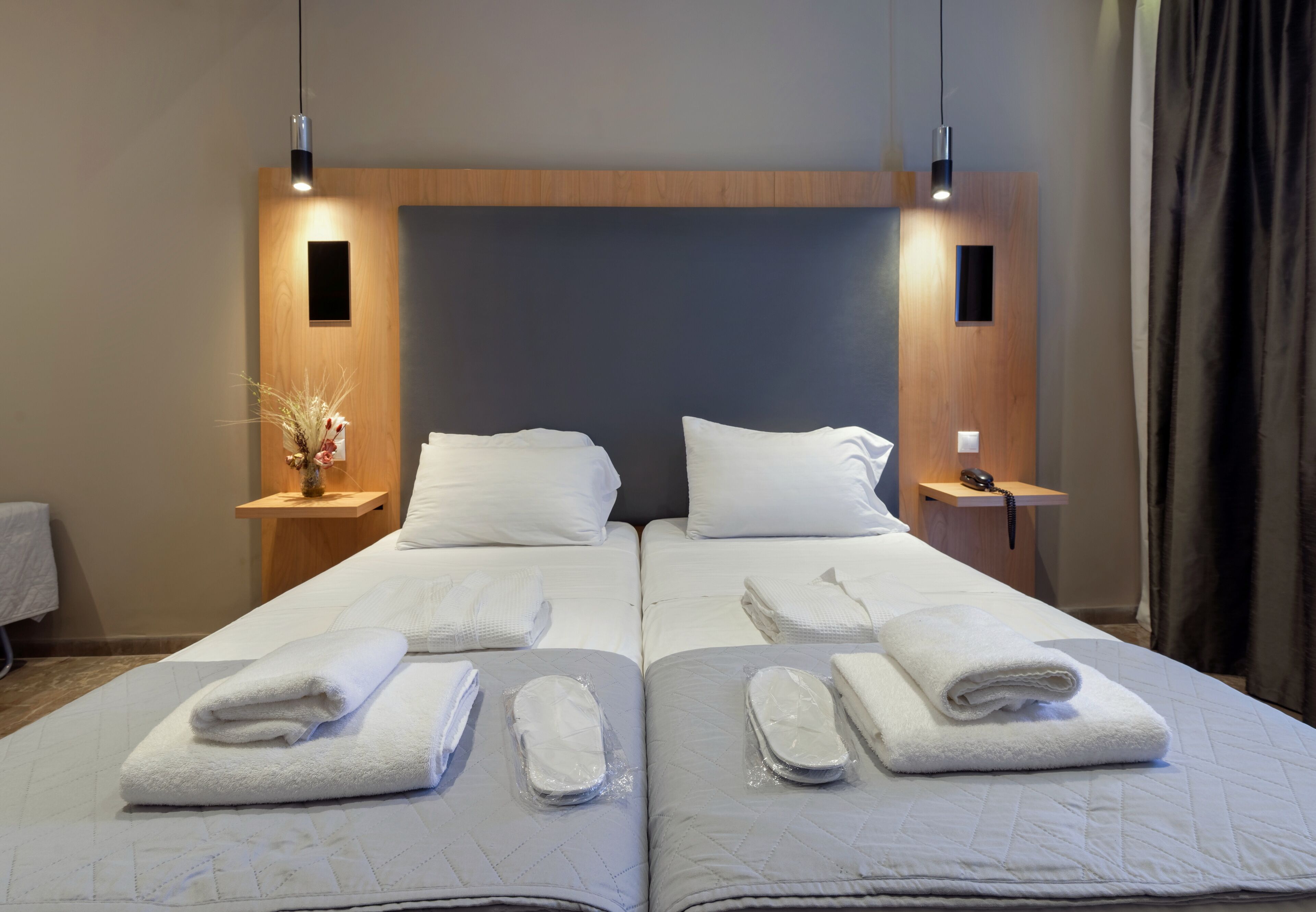 A double bed with white linens and towels, a wooden headboard, bedside tables with lamps, and a vase with dried flowers.