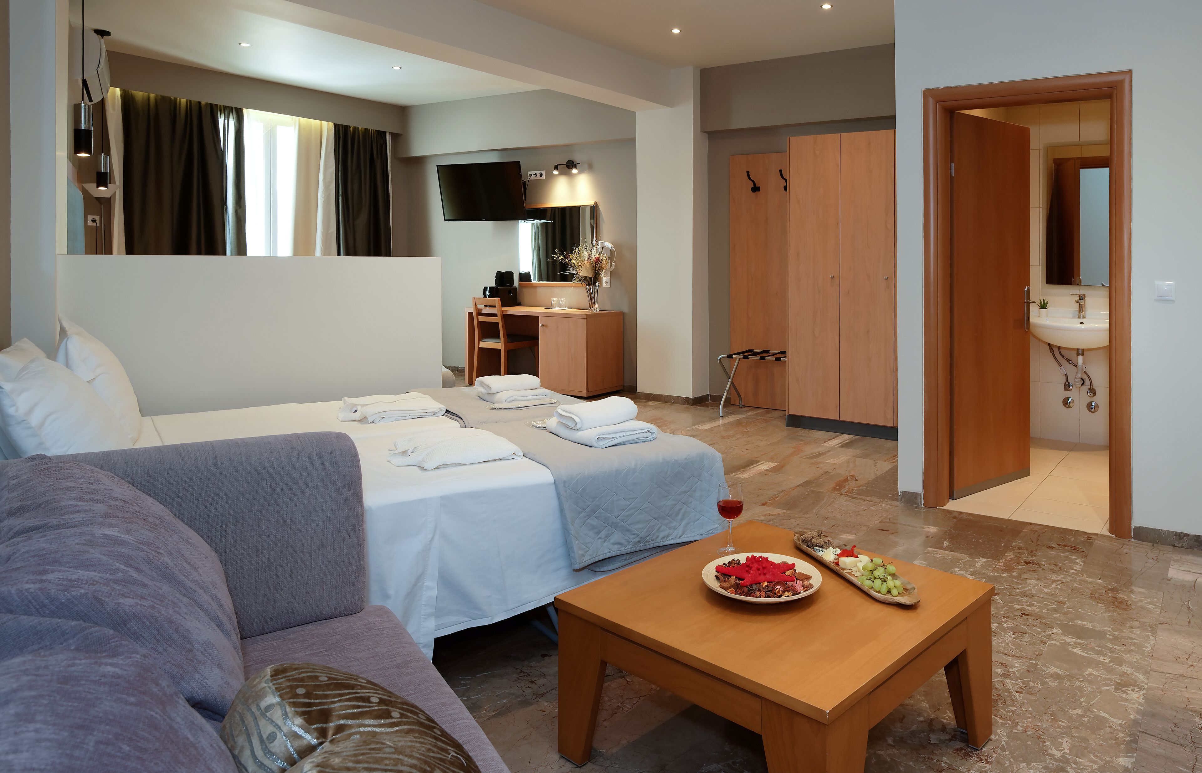 Habitación de hotel con cama, sofá, escritorio con silla, televisor, espejo, armario de madera y baño con lavamanos y espejo.