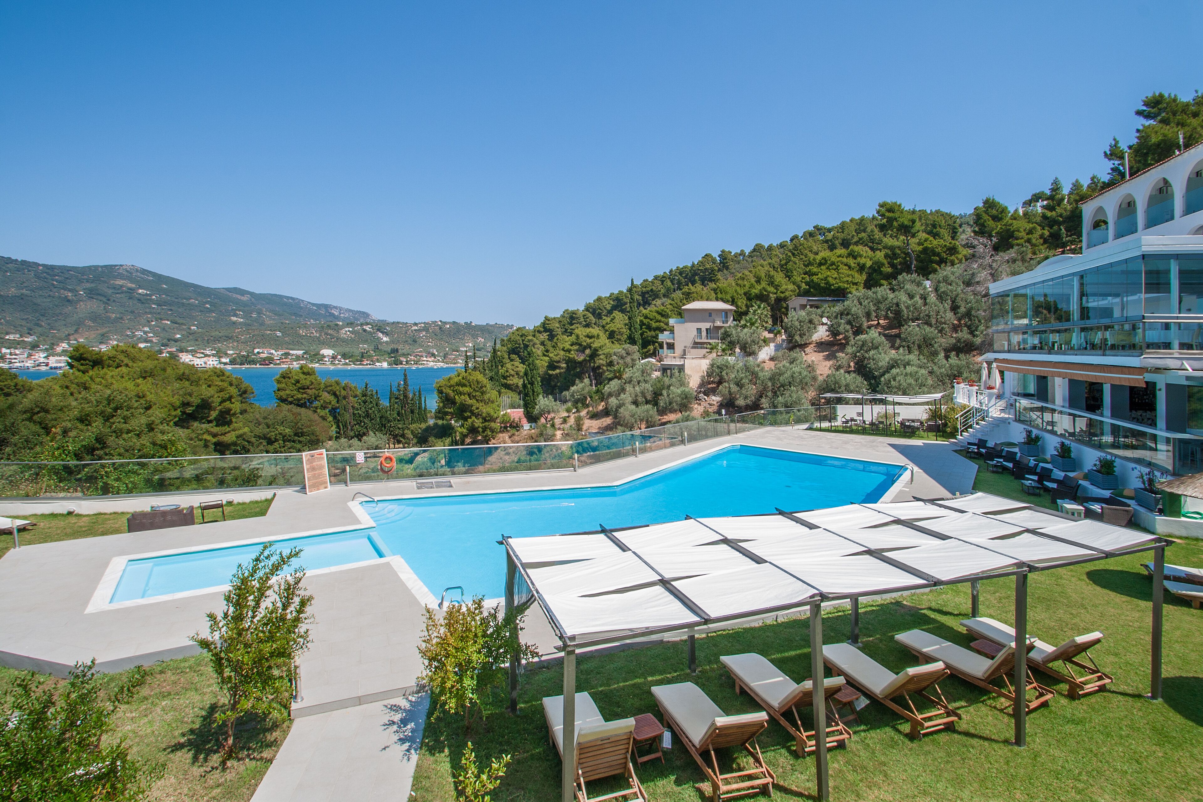 A swimming pool with sun loungers and a covered area, surrounded by greenery and overlooking a coastal town.