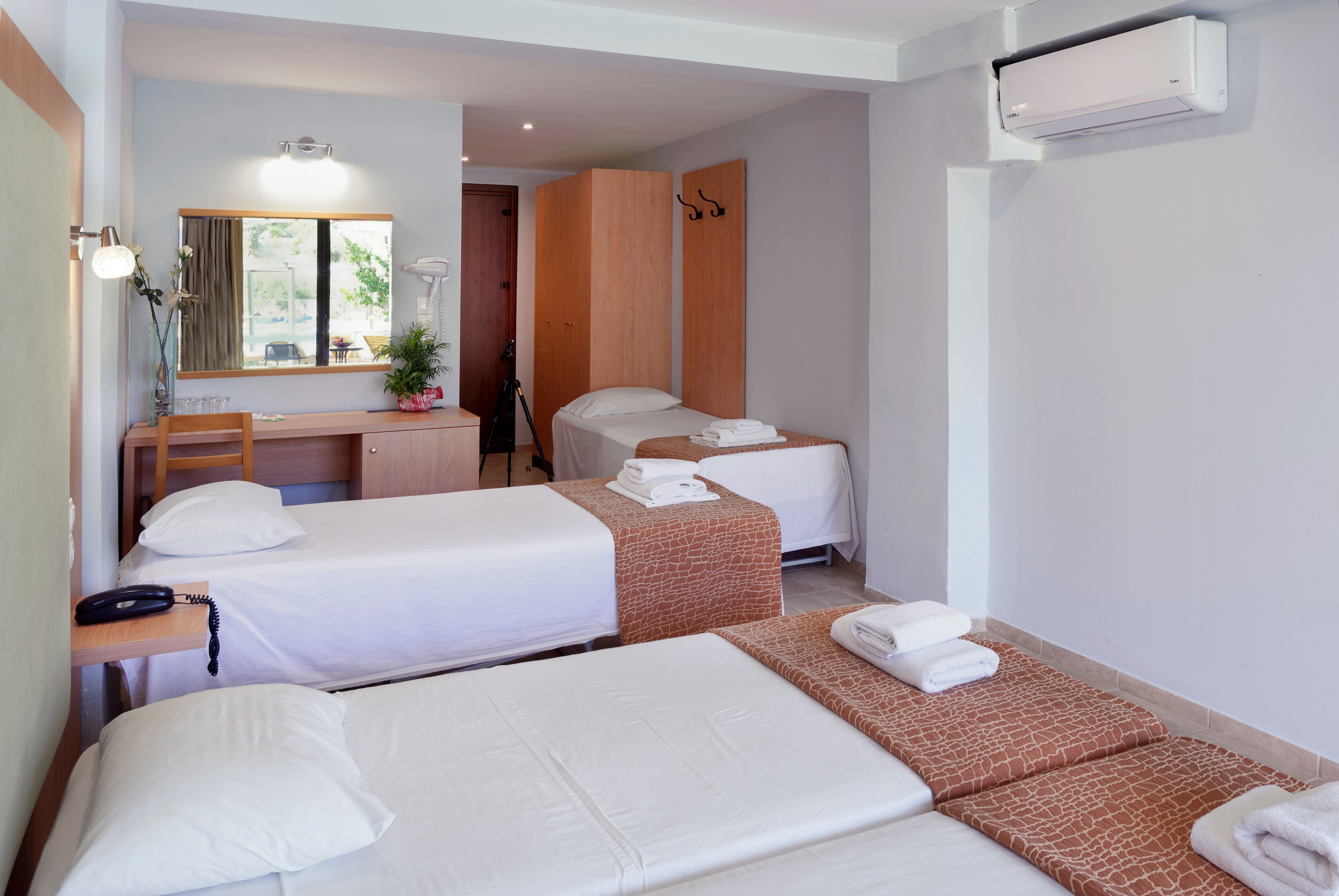 A hotel room with two single beds, a desk with a chair, a mirror, and an air conditioning unit.