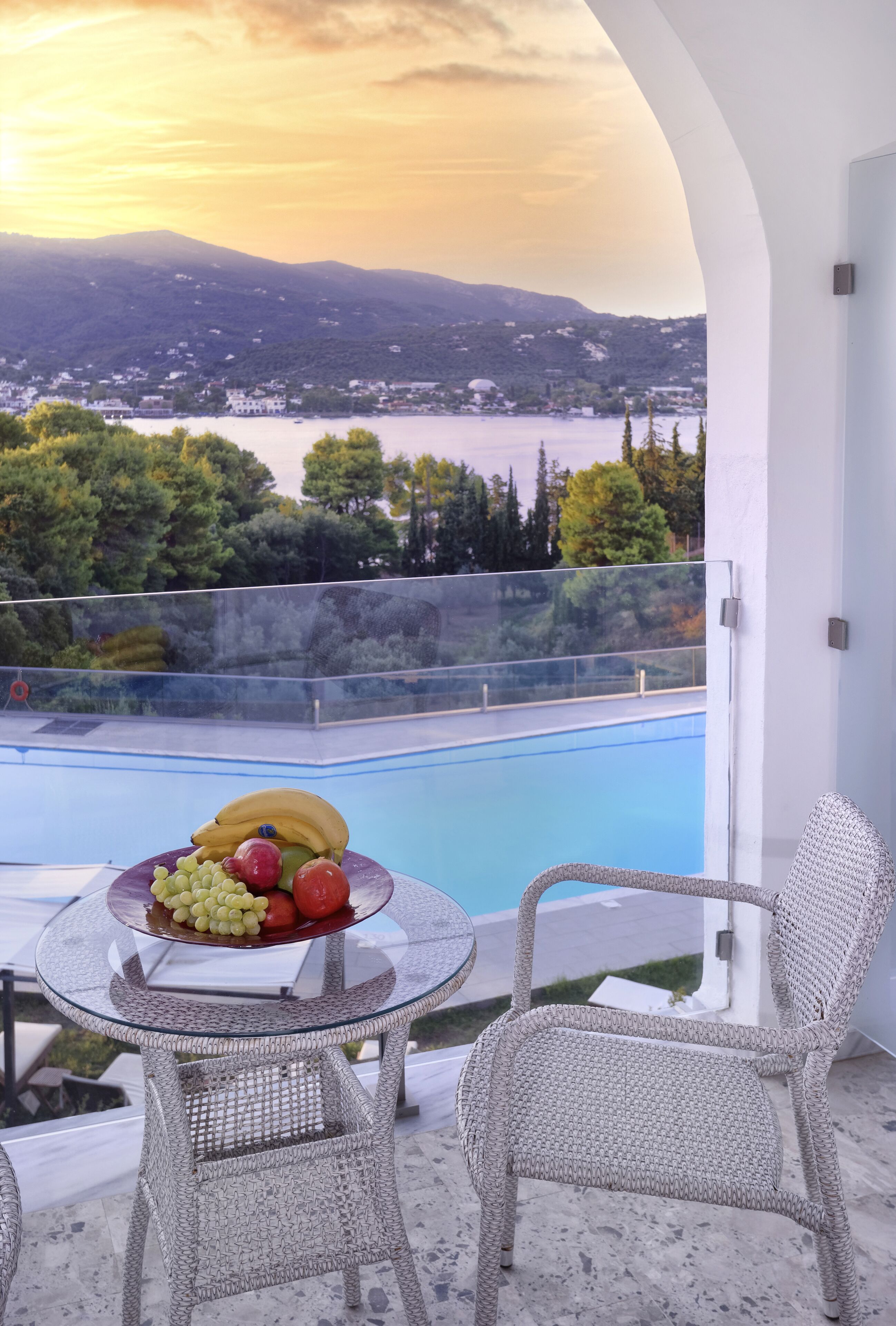 A balcony with a glass table holding fruit, overlooking a swimming pool and a mountainous landscape.