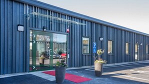 A modern building with a metal exterior, a glass entrance, and a red carpet leading to the door. There are two large planters with flowers near the entrance. - Hotell Eken (Kållered)