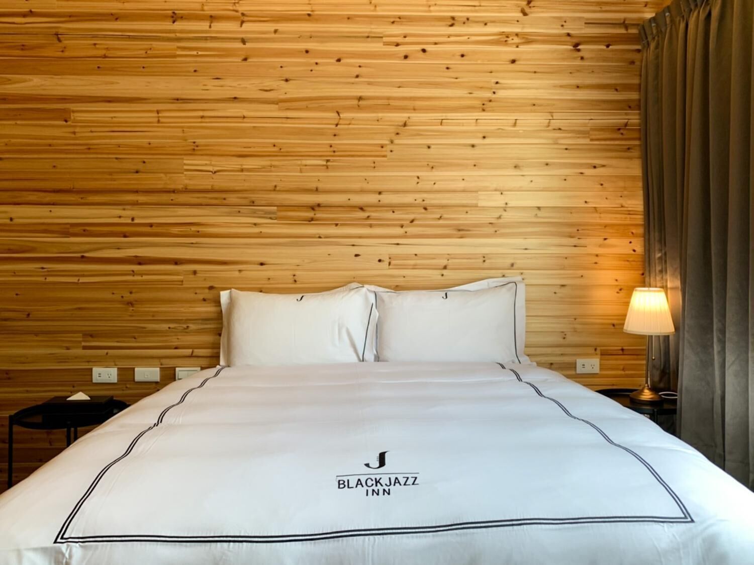 Habitación de hotel con pared de paneles de madera, cama con ropa blanca y lámpara en una mesita de noche.