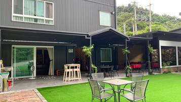 Un edificio moderno de dos plantas con un patio cubierto que incluye una mesa y sillas, rodeado de césped artificial.
