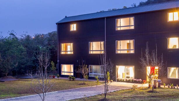 A modern multi-story building with multiple illuminated windows, a gravel driveway, and a small garden area with potted plants. - BlackJazz Inn (Puli)