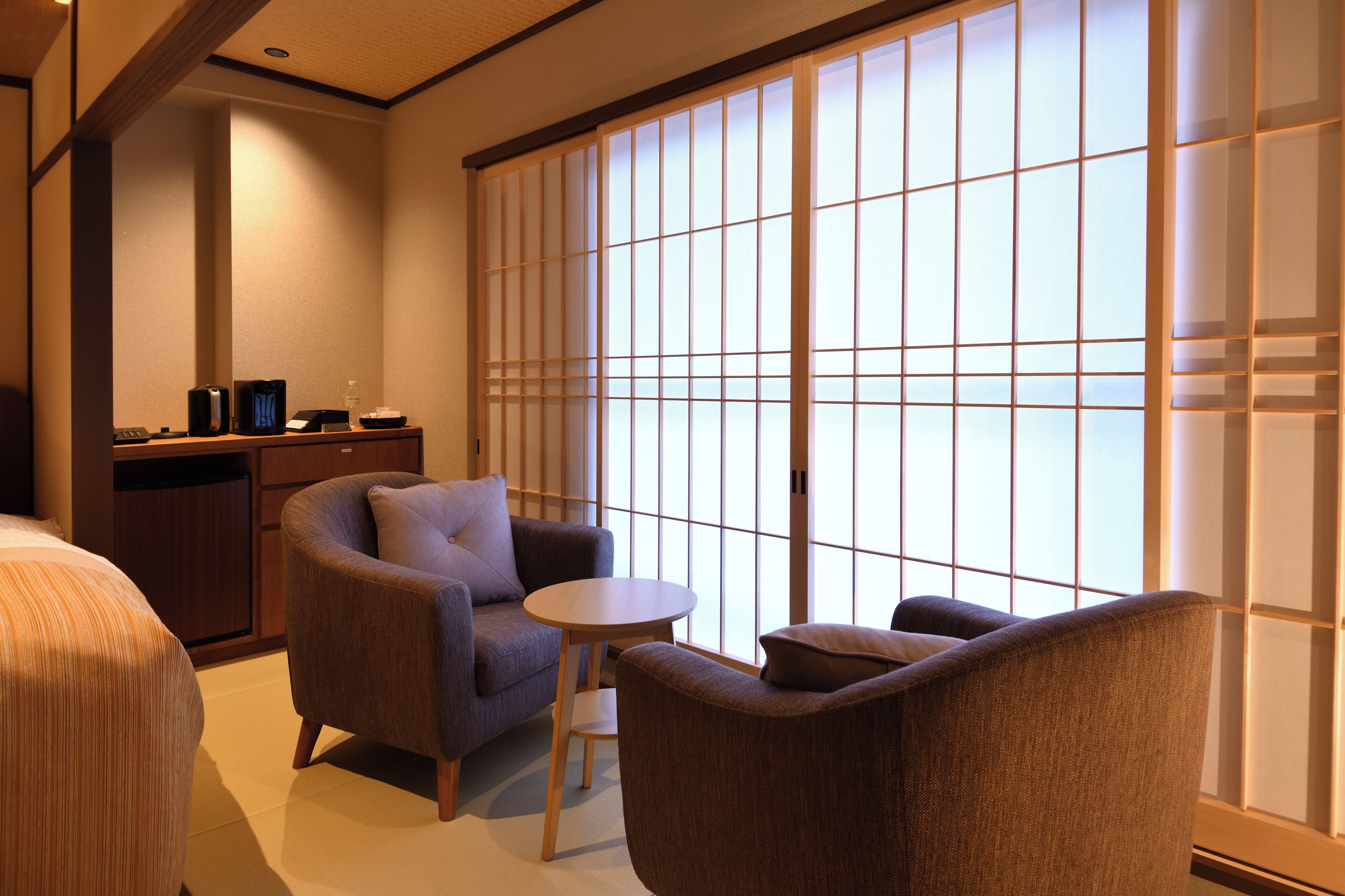 A hotel room with a traditional Japanese-style sliding door, a sofa, a small round table, and a desk with a computer and a lamp.