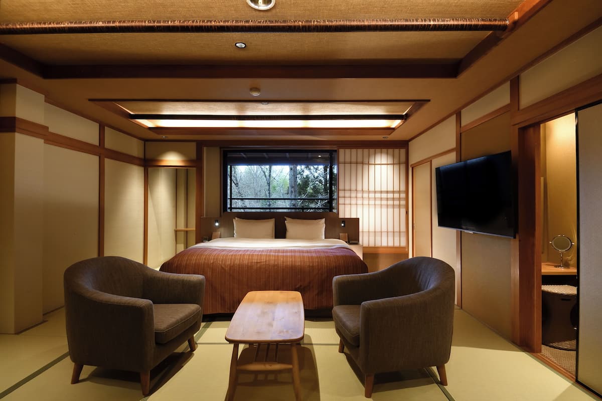 A hotel room with a large bed, two armchairs, a small table, a TV, and a window with a view of trees.