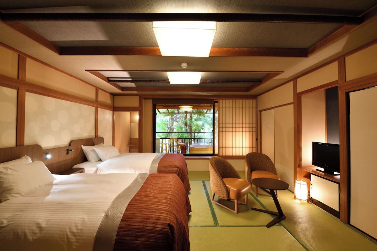 A hotel room with two beds, a television, a chair, and a window with a view of greenery.