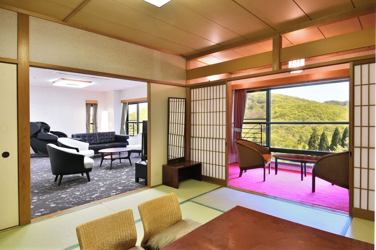 A traditional Japanese room with tatami flooring, a low table, and a view of a lush green hillside through large windows.