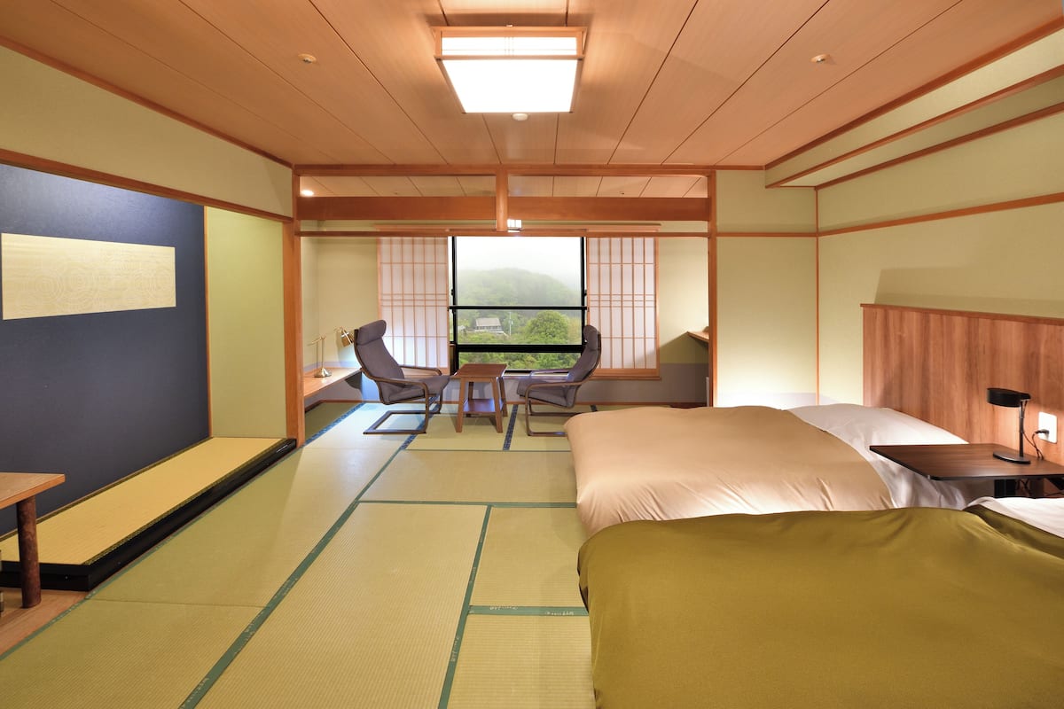 A traditional Japanese-style room with tatami flooring, a bed, a desk, and a view of a mountain through a window.