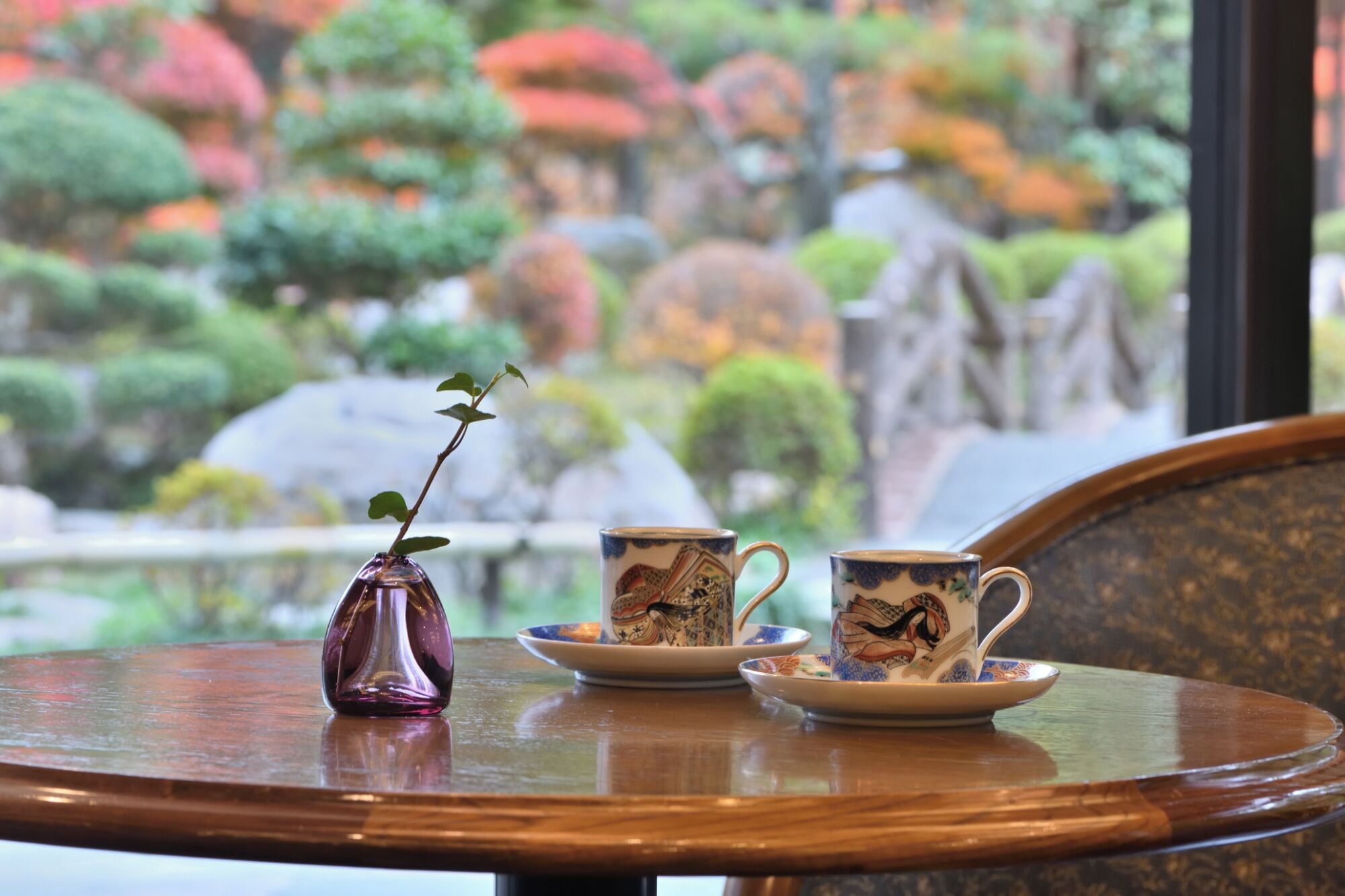 A table with a vase and two cups with saucers, set against a view of a garden with various trees and rocks.