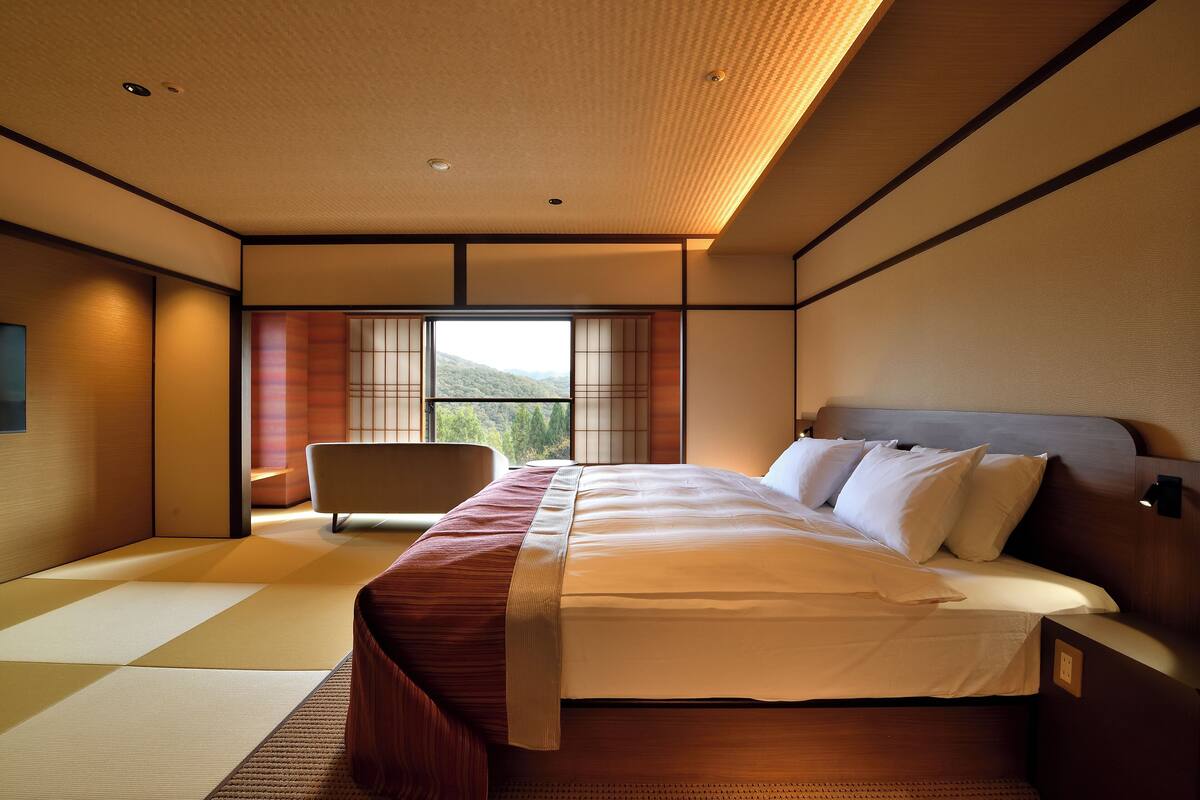 A modern hotel room with a large bed, a sitting area with a chair, and a view of the outdoors through large windows.